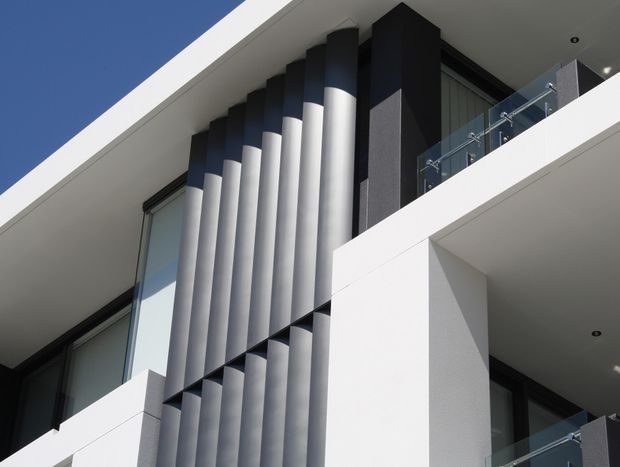 Close-up upward view of a contemporary multi-level building facade. White projecting slabs and walls frame dark glazing, while a strong vertical screen of alternating cylindrical silver and dark elements creates a sculptural architectural feature. Glass balustrades line the upper balconies, and recessed ceiling lights are visible beneath the overhangs against a clear blue sky.