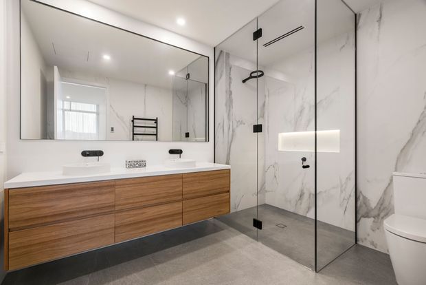 Contemporary bathroom featuring a floating timber vanity with two white basins, a large wall-to-wall mirror, a frameless glass walk-in shower, marble-look wall panels, a grey floor, and a partially visible toilet. The room is bright, clean-lined, and refined, with integrated lighting and minimal black hardware.