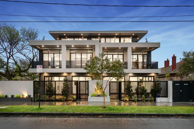 Street-facing view of a contemporary multi-storey residential building with a strong horizontal composition, flat projecting roof slabs, expansive glazed openings, white rendered walls, dark metal privacy fencing and landscaped front entries. Mature trees and neighbouring homes are visible around the property.