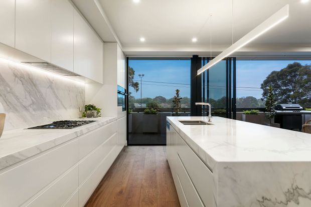 A bright contemporary kitchen with a long white island featuring an integrated sink and waterfall stone end panel. White handleless cabinetry lines the opposite wall beneath a marble-look splashback, with a built-in cooktop and under-cabinet lighting. Timber flooring adds warmth, while full-height black-framed glazing opens to an outdoor barbecue area and leafy suburban outlook. A slim linear pendant runs above the island.
