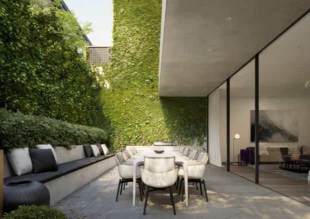 A refined courtyard terrace beside a contemporary glass-walled residence. The outdoor dining setting includes a light stone or concrete table with upholstered chairs and a long built-in bench, surrounded by dense planting and a dramatic ivy-covered wall. Floor-to-ceiling glazing reveals a minimalist living room inside, while a broad concrete soffit creates a sheltered outdoor zone.