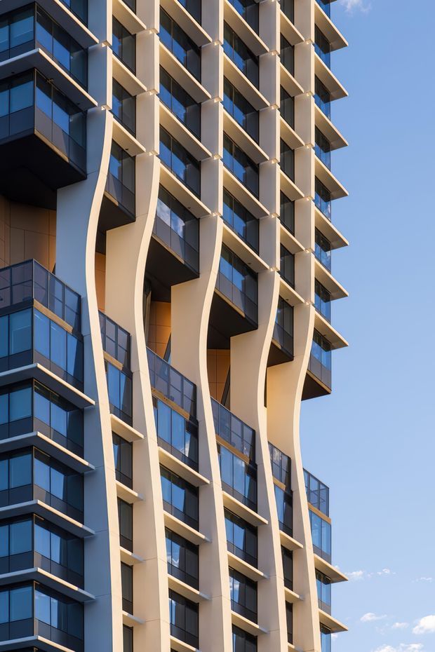 A close-up exterior view of a tall contemporary multi-storey apartment building. The facade features repeated projecting balconies, dark blue reflective glazing, pale sculptural curved vertical columns, and strong geometric horizontal and vertical lines. The building is photographed against a clear blue sky in warm sunlight, emphasizing its modern facade composition.
