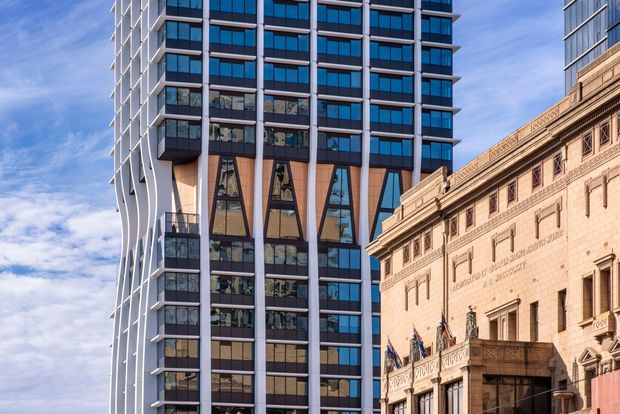 Urban architectural exterior showing a tall contemporary high-rise clad in dark glazing, pale vertical structural fins, and warm tan panels, positioned beside an ornate historic masonry building with classical detailing and decorative parapets. The image highlights the contrast between modern and heritage commercial architecture under a bright blue sky.