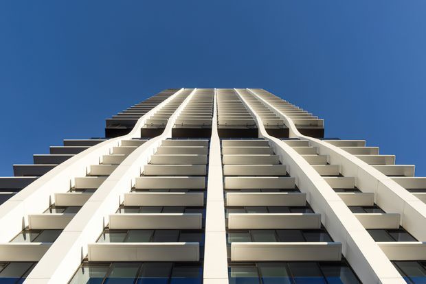 Upward-looking view of a tall modern high-rise facade against a clear blue sky. Repetitive stacked balconies and dark glazed openings are framed by strong white vertical structural elements, creating pronounced symmetry and converging lines.