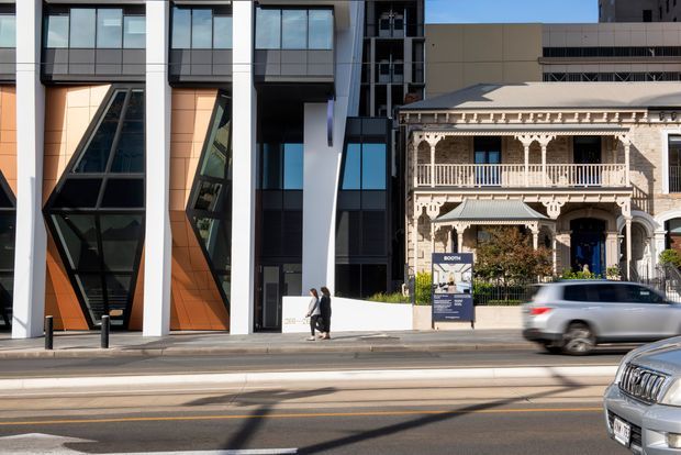 Street-level view of a contemporary multi-storey building with bold white structural fins, dark metal framing, extensive glazing, and orange feature panels beside a restored ornate Victorian-era terrace or mansion with decorative balconies and lacework. The image is captured from across an urban street, with passing vehicles in the foreground.