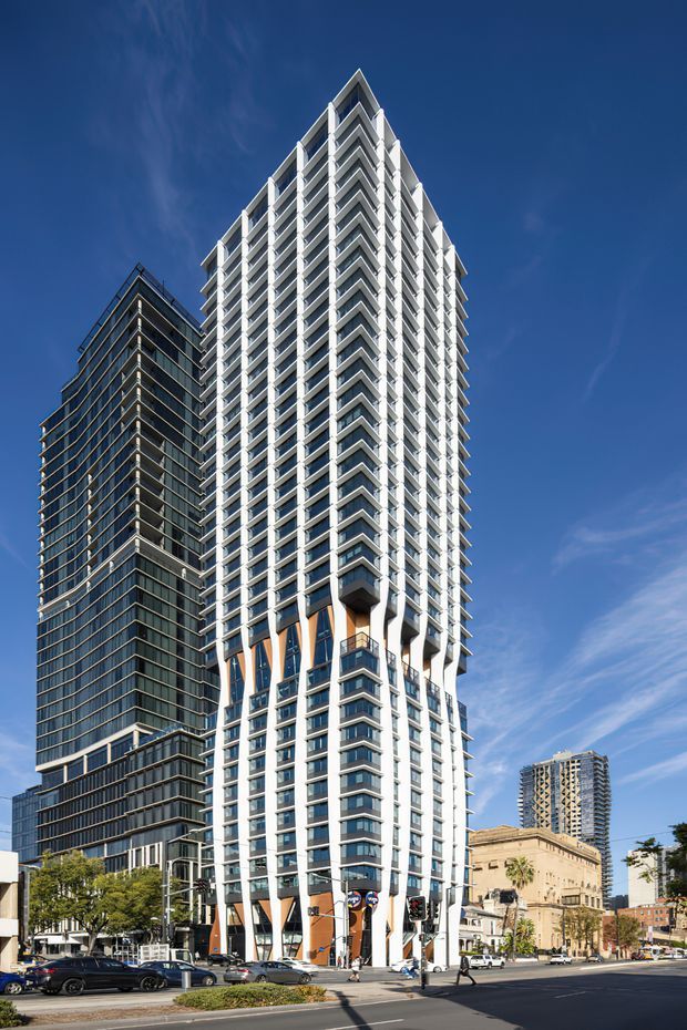 Street-level exterior view of a tall contemporary high-rise residential or mixed-use tower in a dense urban setting. The prominent white tower has a sharply pointed roofline, extensive repetitive balconies, projecting geometric balcony edges, dark glazing, and orange accent panels at the lower levels. A darker glass high-rise stands behind it, with additional city buildings, traffic signals, streets, and vehicles in the foreground under a clear blue sky.