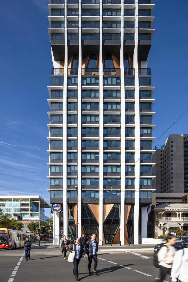 Street-level view of a tall contemporary apartment or mixed-use tower in an urban setting. The facade has repeated projecting balconies, extensive dark glazing, strong vertical white structural fins, and distinctive angular recessed sections with warm timber-toned panels. Commercial or retail frontage and pedestrians occupy the street level, with neighbouring high-rise buildings visible nearby.