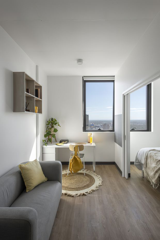 Bright, compact multi-purpose room with a grey sofa in the foreground, a small built-in desk and chair beside a large window, wall-mounted open shelving, and a bedroom visible through a partially open sliding glass door. Light wood-look flooring, white walls and cabinetry, abundant natural light, and a city view create an airy contemporary interior.
