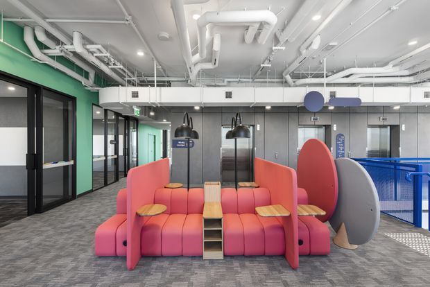 A bright contemporary commercial interior featuring a colourful modular lounge seating installation in an open office or workplace. Coral-pink upholstered seating is arranged around integrated small timber tables and shelving, with curved privacy screens in coral, grey, and blue. The space has exposed white mechanical services and ductwork, dark grey wall panels, extensive black-framed glazed meeting-room partitions, grey carpet tiles, and a blue-railed circulation or stair-edge zone.