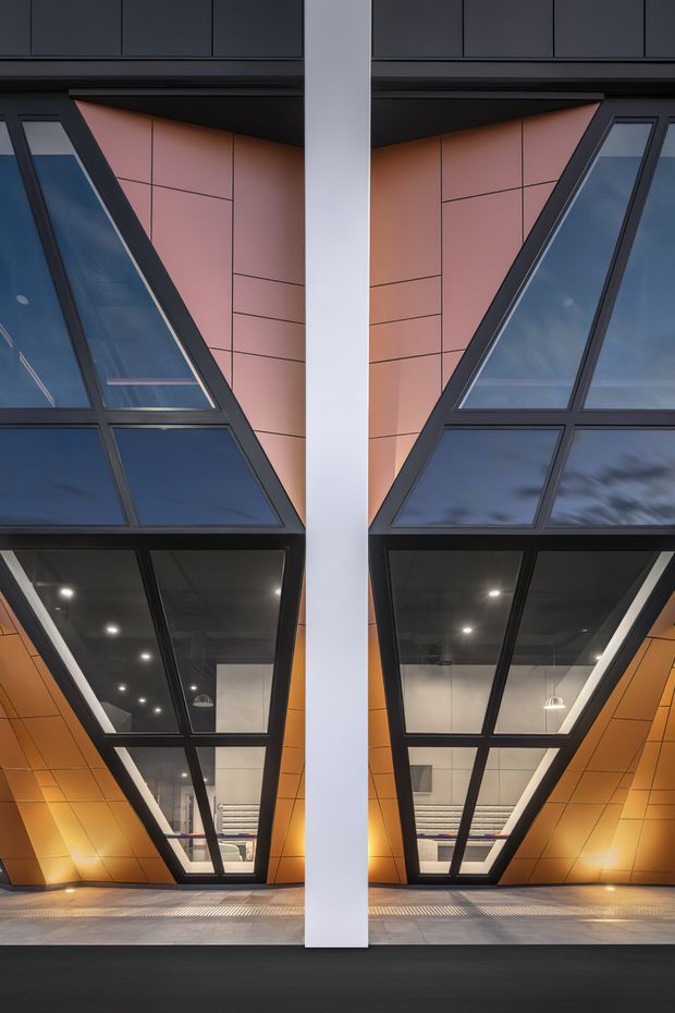 A symmetrical close-up of a contemporary building facade featuring a central white structural column, dark metal-framed angled glazing, and warm orange geometric cladding panels. The reflective glass reveals interior lighting and ceiling fixtures, while the lower portion shows a paved entrance or forecourt.