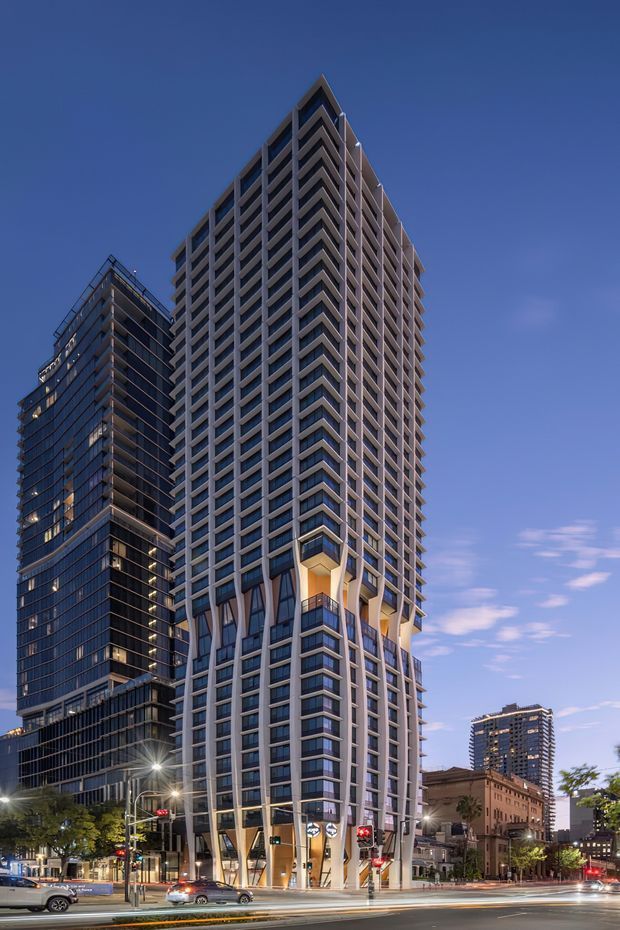 Twilight street-level view of a tall contemporary high-rise tower with a sharply articulated corner, repetitive projecting balconies, extensive glazing, and a light-toned structural grid. The tower rises above a glazed neighboring building and a lower historic urban streetscape, with illuminated ground-floor retail or lobby frontage and active city roads in the foreground.