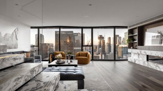 A luxurious contemporary high-rise living room with floor-to-ceiling black-framed glazing overlooking a dense city skyline. The room features a large marble-clad fireplace and matching marble cabinetry, dark timber flooring, a grey sectional sofa with tufted ottoman, sculptural mustard armchairs, recessed ceiling lighting, and restrained contemporary furnishings.
