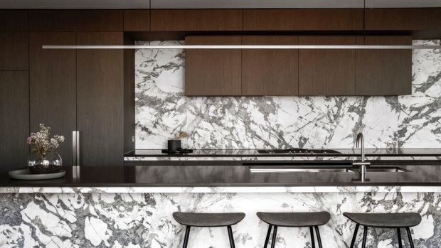 A sophisticated contemporary kitchen with dark timber cabinetry, a dramatic white-and-grey veined stone backsplash and island front, a long linear pendant light, stainless steel fixtures, and three minimalist bar stools at the island.