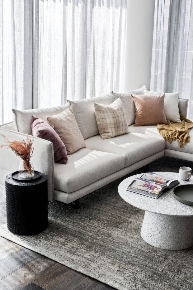 Bright, softly styled living room with a large pale grey sectional sofa positioned beside full-height sheer curtains. Neutral cushions, a textured throw, a dark textured side table, and a round white speckled coffee table create a calm contemporary setting. A large muted grey area rug sits over dark timber flooring, with strong filtered daylight creating striped shadows.