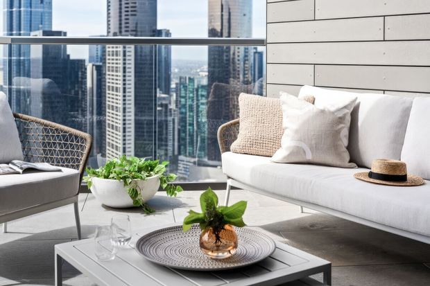 Bright contemporary high-rise living room with floor-to-ceiling glazing overlooking a dense city skyline. The space features a light upholstered sofa, woven lounge chair, pale horizontal wall panelling, a low square coffee table, indoor greenery, and neutral decorative textiles.