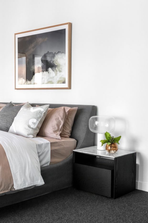 A close-up interior photograph of a refined bedroom corner, featuring a dark upholstered bed with layered white and blush bedding, a black bedside table with a pale stone-like top, a small potted plant beneath a glass lamp, and framed abstract artwork on a white wall. Dark carpet and charcoal furniture contrast with soft neutral textiles.