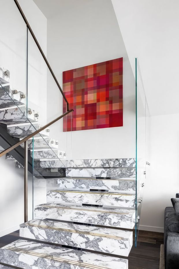 Bright contemporary interior featuring a sculptural open-riser staircase clad in heavily veined white and grey marble, with clear glass balustrades and slim dark metal or timber handrails. A vivid red, orange, and magenta geometric artwork creates a strong feature wall above the stair, while dark timber flooring and part of a grey sofa are visible at the side.
