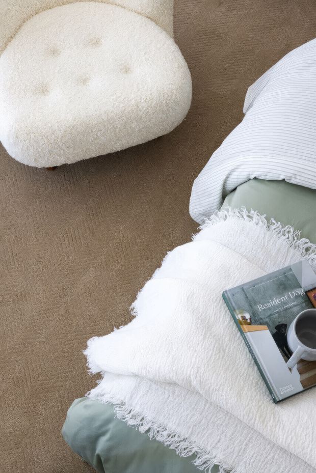 A softly styled bedroom vignette viewed from above, featuring a white textured throw draped over muted sage-green bedding, a striped white pillow, a plush cream rounded ottoman, and a neutral brown carpet. A design book and coffee cup rest on the bed, creating a calm, tactile and inviting atmosphere.