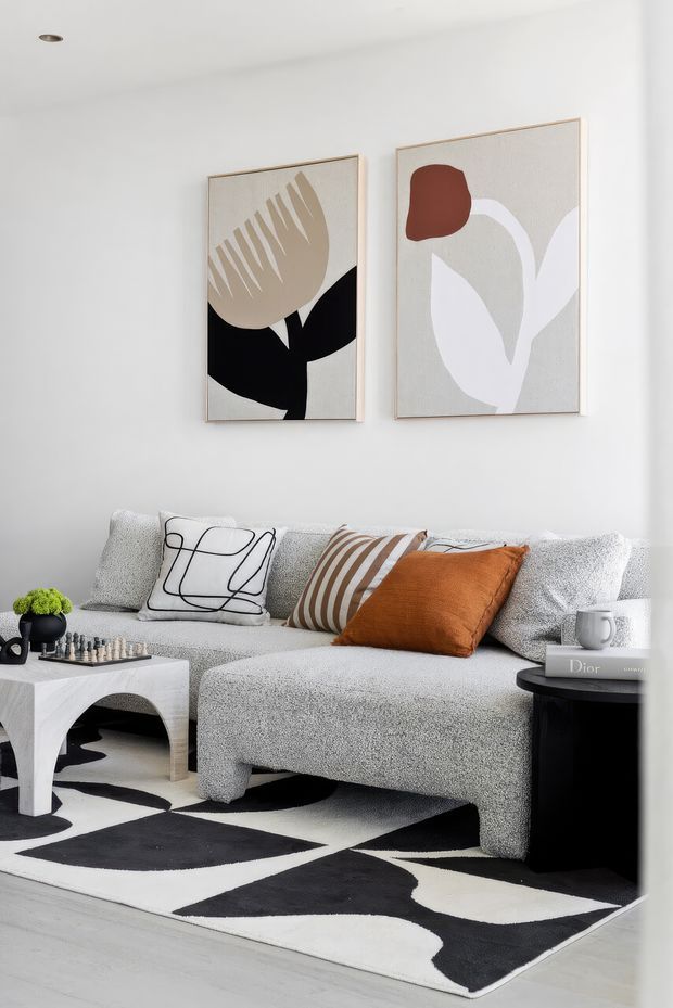 A bright, compact living room with a light textured sectional sofa, graphic black-and-white rug, sculptural white coffee table, and black side table. Abstract botanical artwork hangs on the white wall, while rust-orange and neutral cushions add warmth. A small chess set and potted plant provide decorative detail.