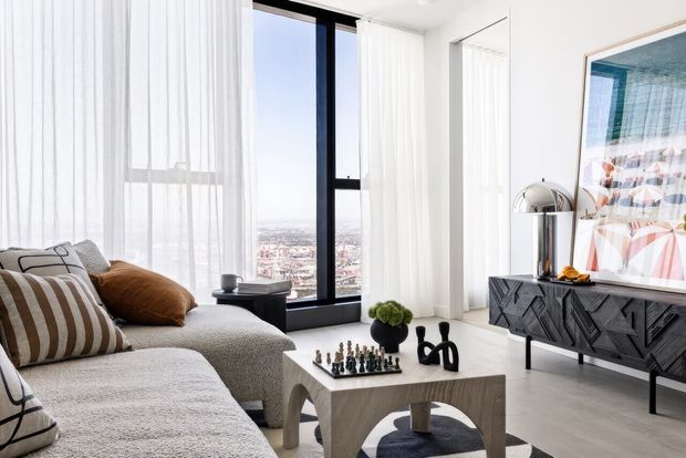 Bright contemporary living room with floor-to-ceiling windows, sheer white curtains, a textured sectional sofa, sculptural wood coffee table, black geometric sideboard, and large abstract artwork. A city view is visible through the glazing.