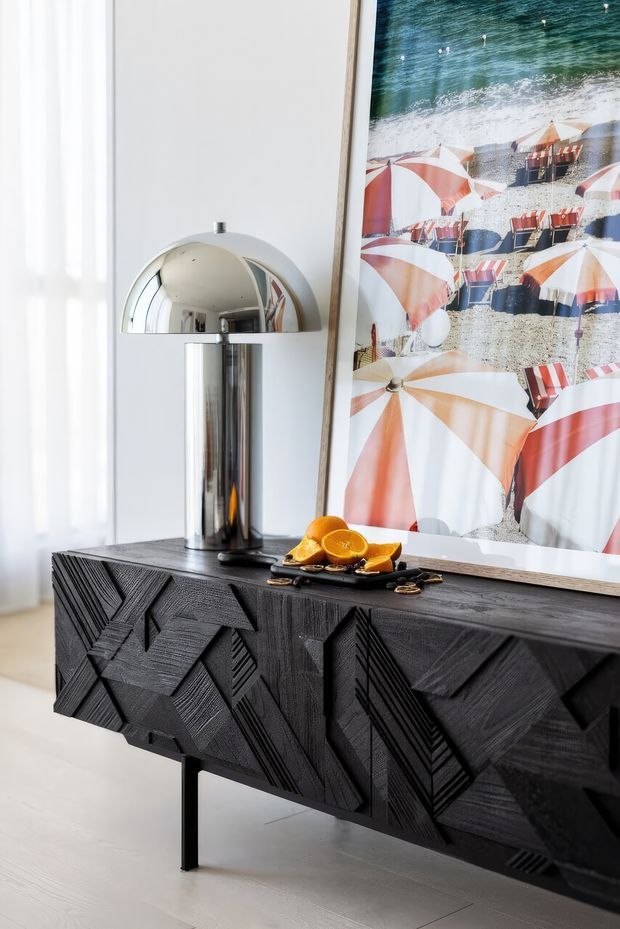 A styled interior vignette featuring a black textured geometric sideboard on slender legs, a polished chrome dome table lamp, and a large framed beach photograph. Decorative oranges sit on a tray atop the sideboard. The bright room has white walls, sheer curtains, and pale flooring.