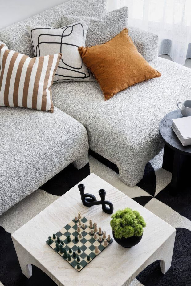 A close-up view of a styled living room featuring a large light-grey textured sectional sofa, layered decorative cushions in white, tan, and brown, and a white stone or timber-look coffee table. The table holds a chessboard, sculptural black decor, and a small potted moss-like plant. A black-and-white geometric rug, dark round side table, and sheer curtained window complete the contemporary setting.