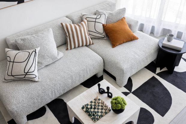 Bright contemporary living room featuring a large light-grey textured sectional sofa, graphic black-and-white patterned rug, white coffee table, sculptural black side table, and cushions in white, striped brown, abstract black linework, and burnt orange. Sheer curtains provide soft natural light.