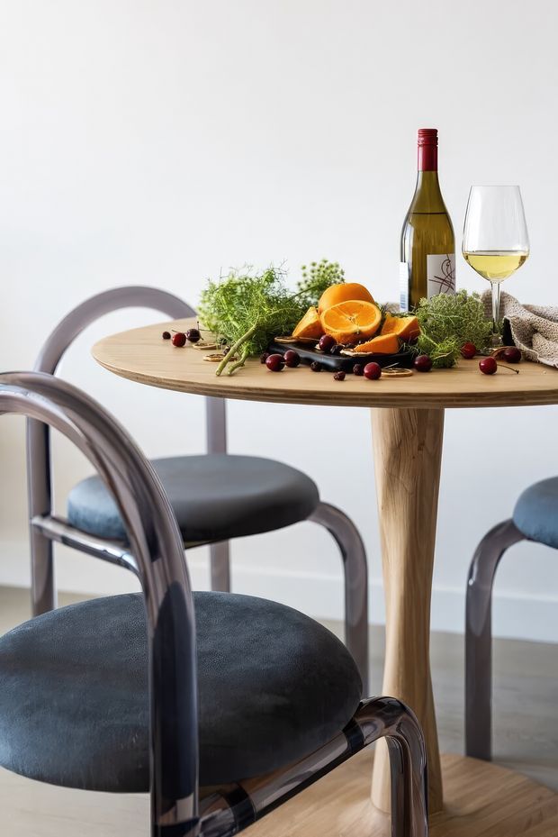 A close-up interior scene of a small round timber dining table with a central pedestal, surrounded by dark upholstered chairs with curved metal frames. The table is styled with a wine bottle and glass, sliced oranges, berries, herbs, and a folded textured napkin against a light wall.