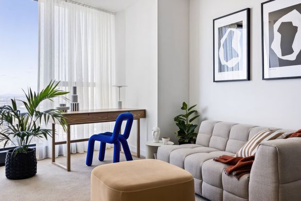 Bright contemporary living room with a large light-grey sectional sofa, tan upholstered ottoman, bold cobalt-blue sculptural chair, indoor plants, and a slim timber console desk beneath abstract framed artwork. Full-height sheer curtains and expansive glazing create an airy, light-filled setting.