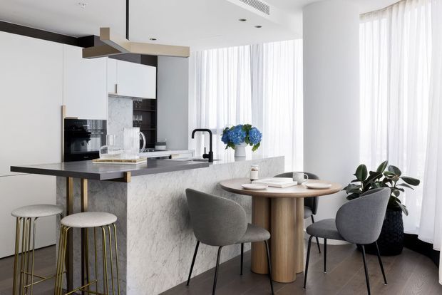 Bright contemporary open-plan kitchen and dining area with a marble-look island, integrated appliances, sculptural pendant light, sheer floor-to-ceiling curtains, and a compact round dining table with upholstered chairs. The palette is predominantly white, grey, black, and warm timber.