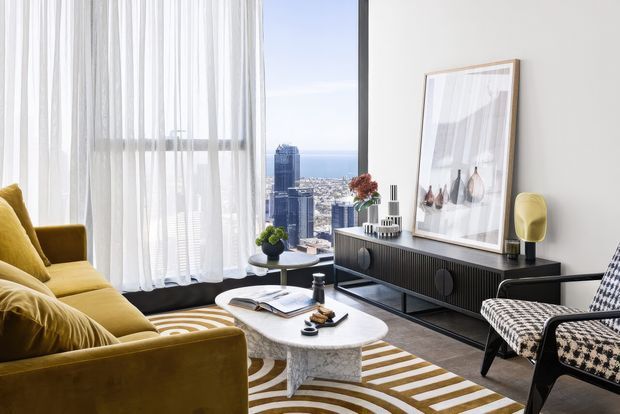 A stylish high-rise living room with floor-to-ceiling windows overlooking a dense city skyline and water beyond. The room features a mustard velvet sofa, a matching lounge chair, a black-framed patterned armchair, a white oval coffee table, a dark ribbed console, sheer white curtains, a large framed artwork, and a bold geometric rug.