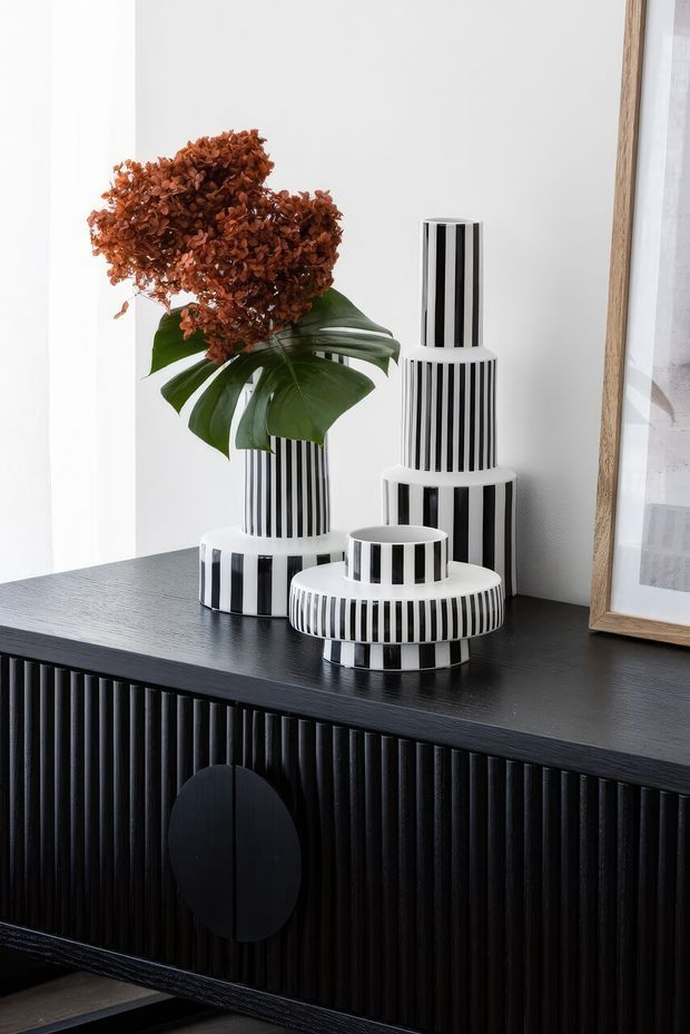 A styled interior vignette featuring a black fluted sideboard with a round door pull, topped with sculptural black-and-white vertically striped ceramic vases. One vase holds dried rust-coloured hydrangea-like foliage and broad green leaves. A framed artwork leans against the wall at right, reinforcing the bold monochrome and warm accent palette.