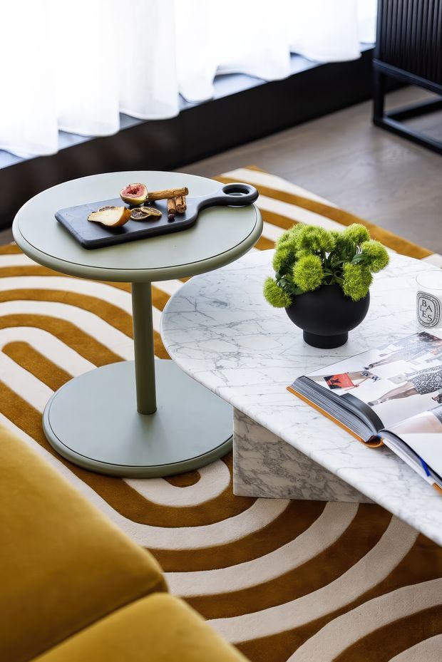 A styled living room vignette featuring two round nesting-style coffee tables, one with a pale green finish and another with a white marble surface and stone base. A black serving board with snacks sits on the smaller table, while a black planter with bright green foliage and an open magazine decorate the marble table. A mustard and cream concentric-patterned rug, mustard upholstered sofa, sheer curtains, and dark wall detailing create a warm, contemporary setting.
