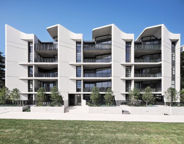 Exterior architectural photograph of a contemporary multi-storey apartment building with three prominent sections, sculptural curved white rendered facade elements, dark recessed balconies, black metal balustrades, extensive glazing, and landscaped raised planters along the frontage. The building is viewed from across a lawn under a clear blue sky.