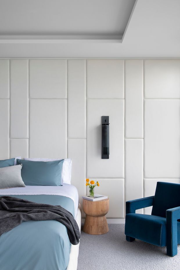 A refined bedroom interior featuring a bed with pale blue bedding, grey cushions and throw, a full-height white padded panelled feature wall, a black vertical wall light, a small rounded timber bedside table with flowers, and a deep blue upholstered armchair. The room has a recessed ceiling detail and soft neutral carpeting, creating a calm, contemporary atmosphere.