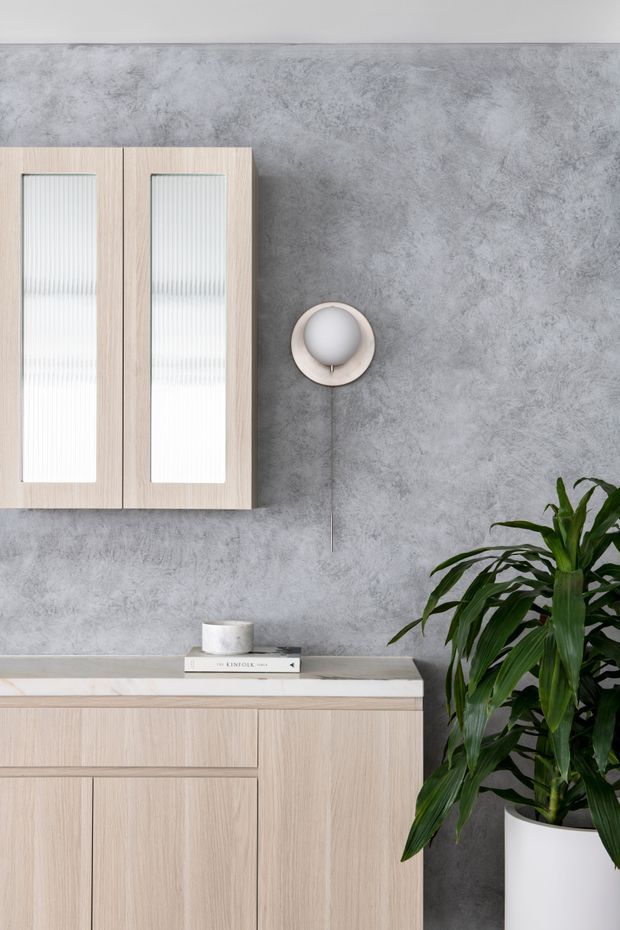 A close-up interior view of a refined storage and display area. Pale timber wall cabinets with vertically ribbed translucent inserts hang above a matching low sideboard with flat-panel doors and drawers. The cabinetry sits against a mottled grey textured wall, with a simple round wall light, a small ceramic vessel, books, and a large leafy plant in a white pot adding understated decoration.