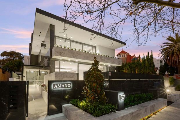 Dusk exterior view of a multi-storey contemporary building with a sharp flat roof, extensive floor-to-ceiling glazing, white rendered walls, dark stone-clad boundary walls, balconies, and a landscaped entrance. A prominent illuminated AMANO sign is visible at the front gate, with neighbouring buildings and mature trees in the background.