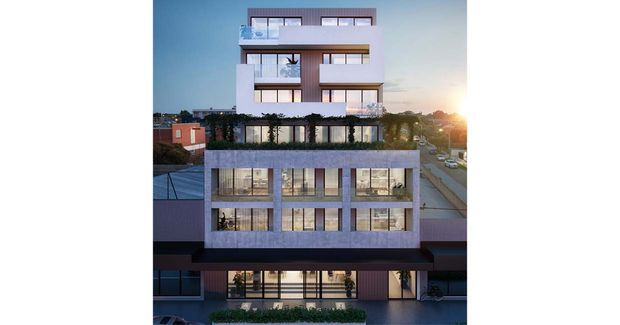 Architectural rendering of a contemporary multi-storey apartment building with a stepped, rectilinear form, extensive glazing, projecting balconies, landscaped rooftop or terrace areas, and a concrete-toned lower facade. The building is shown in an urban streetscape at sunset.