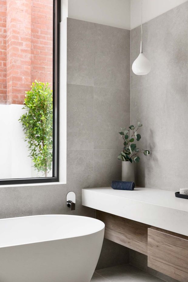 Contemporary bathroom featuring large-format grey stone-look wall tiles, a freestanding white bathtub, floating white vanity with warm wood cabinetry, a black-framed window, and a white pendant light. Natural greenery and exposed brick are visible beyond the window.