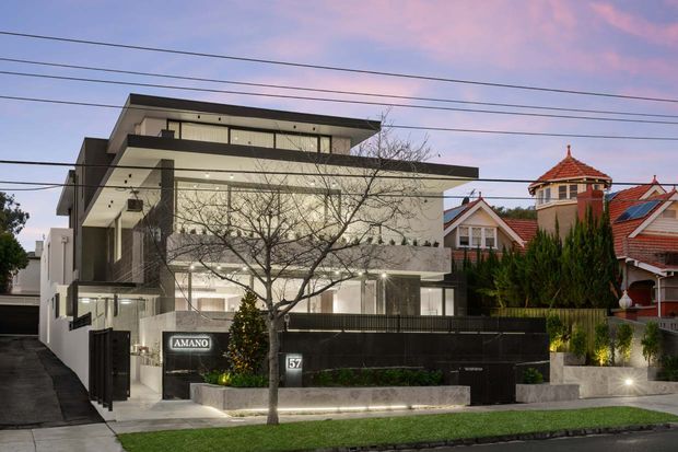 Street-facing view of a contemporary multi-storey residence with a flat roof, projecting upper levels, broad glazing, concrete and dark facade finishes, and a landscaped front boundary. Traditional neighboring homes with red tiled roofs are visible behind it.