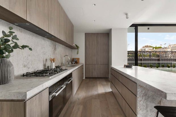 Contemporary galley-style kitchen with extensive warm wood cabinetry, a pale stone benchtop and splashback, integrated stainless-steel appliances, and a central island. Large windows and a glazed door provide natural light and views to an outdoor area, while subtle greenery adds an organic accent.