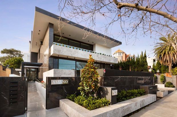 Street-facing view of a contemporary multi-storey residence with a flat roof, projecting upper-level balcony, extensive glazing, dark stone-clad boundary walls, and a landscaped front entry. The facade combines light rendered walls, dark framed windows, and polished dark cladding, with neighboring residential buildings and mature planting visible around it.