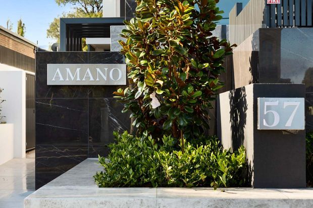 Street-facing entrance to a contemporary property, featuring dark marble-look wall cladding, a bold AMANO sign, modern black metal framing, a large number plaque, and carefully arranged evergreen landscaping. The facade combines dark stone-like surfaces with light rendered walls and a clean, geometric entry design.