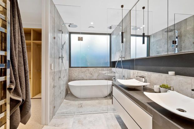 Contemporary bathroom featuring a freestanding white bathtub, frameless glass shower enclosure, double floating vanity with vessel basins, large mirrored wall, and grey stone-look wall and floor finishes. Warm timber shelving and dark accents add contrast to the bright, refined space.