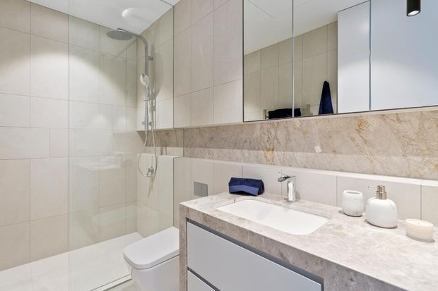 Contemporary bathroom with a frameless glass walk-in shower, wall-mounted shower fittings, toilet, large mirrored wall, and floating vanity with an integrated white basin. Pale large-format wall tiles and a light natural stone vanity surface create a clean, neutral finish.
