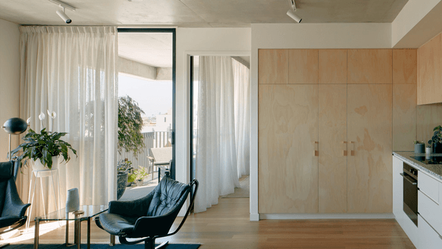 Bright, compact open-plan interior with a living area adjoining a minimalist kitchen. Sheer full-height curtains frame glazed openings, while a black leather lounge chair and sculptural side table sit on pale timber flooring. The kitchen features light plywood cabinetry, white lower units, and a dark stone benchtop. Track lighting and restrained detailing create a clean, contemporary atmosphere.