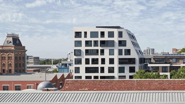 Urban architectural exterior view featuring a contemporary multi-storey apartment building with an irregular white facade, varied dark-framed windows, recessed balconies, and a sloping side elevation. Historic brick and ornate masonry buildings, corrugated metal roofing, and additional apartment buildings are visible in the surrounding city context beneath a partly cloudy sky.