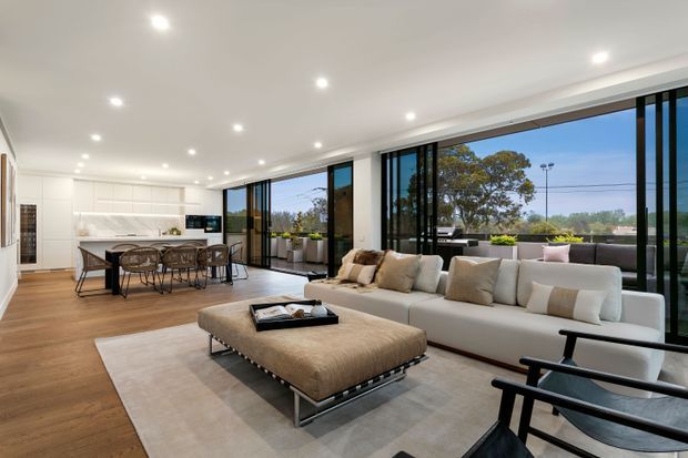 Open-plan contemporary living room with a large cream sectional sofa, oversized upholstered ottoman, and black-framed floor-to-ceiling glazing. A dining area and streamlined white kitchen with island are visible in the background. Warm timber flooring, a pale rug, recessed ceiling lighting, and indoor planter boxes create a bright, refined interior with strong indoor-outdoor connection.