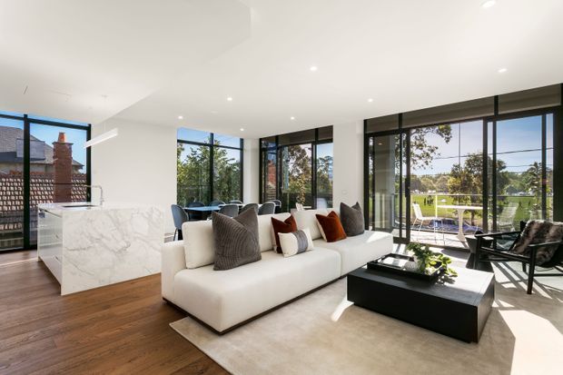 Bright open-plan contemporary living space with a large white sectional sofa, black rectangular coffee table, warm timber flooring, and a white marble-look kitchen island. Floor-to-ceiling black-framed glazing opens the room to a landscaped outdoor area and pool, while recessed ceiling lights and minimal furnishings create a clean, spacious feel.