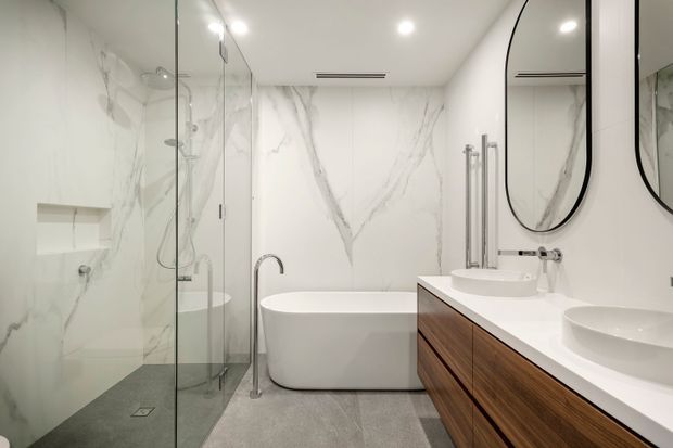 A bright, contemporary bathroom featuring a walk-in glass shower on the left, a freestanding white bathtub, a double vanity with warm wood cabinetry and white benchtop, large white marble-look wall surfaces, grey floor tiles, and rounded black-framed mirrors.